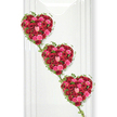 Three intricately crafted rose hearts descend in a gentle diagonal across a spotless white background, creating a romantic and dignified focal point suited to a funeral or memorial service. Each heart is entirely composed of fresh roses in rich shades of red and pink, the blossoms packed so closely that the surface looks soft and plush to the touch. Deep crimson roses dominate, lending depth and gravitas, while clusters of hot pink and softer blush blooms add warmth and an air of affectionate remembrance. Petals layer over one another in tight spirals and open rosettes, giving each heart a textured, almost sculptural quality. Around the perimeter of every heart, delicate ivy winds in a continuous, looping outline; its slim stems trace the contours and its small, pointed leaves dot the edge with refreshing green. These ivy trails stretch from one heart to the next, linking them visually and symbolically, as if joining the stories of those being remembered. The background is a crisp white wall, smooth with faint vertical joins that suggest a panelled interior, much like the serene spaces around Highgate where families gather for farewells, near local churches or overlooking the slopes by Highgate Cemetery. Even, cool-toned light bathes the arrangement, enhancing the intensity of the red and pink hues while casting soft shadows that quietly define the forms. The overall impression is of enduring love and connection, a floral artwork that honours a life with beauty and tenderness. It reflects the compassionate craftsmanship of Highgate florists who specialise in creating personal, heartfelt tributes for the local community.