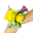 A compact wrist corsage rests diagonally across a light-to-medium forearm, photographed against a crisp white background so every colour and texture reads clearly. Two buttery yellow roses form the joyful centre-one fully open on the left, the other a slightly cupped bud to its right-each petal layered with the soft, velvety texture of fresh garden roses and each heart punctuated by a tiny, sparkling clear rhinestone that catches the studio light. Rising gently behind the roses is a single slender calla lily in a deep plum-to-black-cherry tone; its smooth, trumpet-shaped curve offers a dramatic silhouette and a cool, almost satiny sheen that contrasts with the warm, fragrant roses. Around the blooms a halo of fine asparagus-fern adds airy, feathery movement while broader variegated leaves-pale-edged pittosporum or similar foliage-create a sturdy, graphic base that keeps the corsage balanced on the wrist. Scattered clusters of minute white filler flowers tuck into the design, like tiny stars that soften the composition and add fullness without weight. The floral materials suggest a light, citrus-rose scent with a whisper of green; the rhinestones lend a celebratory glint suited to prom, wedding, or a formal evening. The arrangement is neatly bound so the band itself is mostly hidden, presenting a wearable piece that reads as both timeless and modern. As a local touch, this is the sort of corsage a Highgate bride or a guest walking up Southwood Lane might choose to complement warm evening light and classic tailoring.