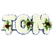 The image shows a personalised funeral flower tribute of the name TOM designed in three bold, upright capital letters, a style often chosen by families in Highgate to create a truly individual farewell. Each letter is fully covered in a dense layer of small white chrysanthemums, arranged closely together so that their overlapping petals form a smooth, even cushion of white. This clean, snowy surface provides a sense of peace and simplicity, allowing the eye to rest gently on the name. Around the outer edge of each letter, a royal blue satin ribbon runs in a neat, pleated band, outlining the shapes and giving them a formal, polished look. The blue makes the letters stand out clearly and adds a traditional accent that feels appropriate for a dignified service near Highgate's tranquil green spaces. At specific points on the tribute-just above the left arm of the T, along the curve of the O, and on the upper right corner of the M-the florist has placed small, carefully balanced clusters of accent flowers. These feature rich, deep red roses, smaller cream and white spray roses, and slender white freesia-like blooms, all surrounded by fresh green foliage and soft fern fronds. The red and green against the white chrysanthemums introduce warmth and emotion, symbolising love, memory, and life. All three letters are mounted side by side on a slender dark frame, creating a straight horizontal display ready for a hearse, chapel, or graveside setting. The tribute feels intimate and respectful, a thoughtfully crafted floral arrangement paying quiet homage to Tom within the calm, reflective character of Highgate.