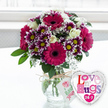 The photograph shows a joyful floral-and-balloon gift set arranged on a pristine white tabletop, bathed in soft daylight that could easily belong to a Highgate flat overlooking Waterlow Park. A rounded clear glass vase stands at the centre, filled with water that magnifies the tidy bundle of green stems gathered inside, each one carefully cut and positioned. Above the vase, the bouquet forms a lush, rounded shape alive with colour. Several large gerbera daisies in an intense magenta-fuchsia hue dominate the design, their broad petals circling dark, velvety centres that add drama and depth. Clustered nearby are smaller chrysanthemums with many narrow petals, each blossom dyed a rich, royal purple that fades into bright white at the tips, cradling cheerful yellow centres that catch the light. Creamy, almost ivory-white roses are interspersed throughout, their gently swirling petals still mostly unfurled, contributing a classic, romantic softness. Wispy sprays of white baby's breath drift between the larger blooms like tiny stars, giving the bouquet a delicate, lacy texture, while fresh green foliage peeks out at the edges, enhancing the natural, freshly picked feel associated with local flower delivery from Flowers Highgate. The neck of the vase is wrapped with a piece of rustic natural twine tied into a modest bow, supporting a small white gift tag printed with a stylised pink rose and green script, suggesting care and craftsmanship. To the right of the vase, slightly overlapping it, rests a shiny, heart-shaped foil balloon. Its silver border gleams, and its white centre features the phrase Love n' Hugs 24-7 in playful, multicoloured letters patterned in pink, purple and blue. The background is softly blurred, likely a bright window, drawing all attention to this affectionate arrangement, ideal for expressing love, gratitude or encouragement to someone special in Highgate.
