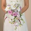 In a tender moment captured within the refined ambience of Highgate, this image focuses on a bride holding an extravagant, cascading bouquet that becomes a visual ode to spring and romance. Large, shimmering Phalaenopsis orchids in pristine white draw the eye, their golden-yellow centres offering a quiet luminescence. Clusters of blush pink and lavender roses, ranging from demurely furled buds to voluptuous open blooms, create sumptuous layers amid the sweep of the arrangement. White baby's breath, airy and light, catches on the edges, bringing a gentle cloud-like softness, while strands of magenta amaranthus trace bold, velvety lines down the bouquet's length. Sprinkles of lavender blossoms and bright green stems flicker in the natural light, playing off the rich foliage that peeks through. The bride, semi-visible in an ivory dress accented with delicate lace, stands before a softly neutral backdrop that ensures all emphasis lands on the artistry of the bouquet. Floral fragrances suggest nearby Highgate Woods in spring; the design, at once structured and fluid, epitomises the balance of tradition and contemporary spirit sought after by North London couples planning their perfect wedding ceremony or reception.