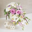 A refined floral arrangement designed as a wedding or celebration table centerpiece is presented in a close, intimate view, as if already placed on a linen-covered table in a Highgate venue. The flowers are artfully gathered in a low, white wicker basket, whose rounded shape and fine weave offer a subtle rustic foundation for the otherwise luxurious blooms. At the heart of the composition, pastel pink roses are open to full perfection, their layered petals forming deep spirals that speak of tenderness and enduring affection. Woven through these roses are stems of white Phalaenopsis orchids, each blossom displaying broad, silky petals and a warm yellow centre, introducing an exotic grace that feels at once modern and timeless, like something you might see at a chic gathering near Highgate Woods. Tucked among the main flowers are white Lisianthus blooms, some with delicate lavender-purple edging, which add a whisper of colour variation and a soft, romantic character. Pale pink stock flowers provide both gentle height and a softly tumbling effect over the basket's rim, their numerous small florets lending a touch of informal charm to the otherwise polished design. Sprinkled throughout, tiny white baby's breath creates airy clusters that float between the larger blooms, unifying the palette and giving the arrangement a light, cloud-like softness. Lush green foliage peeks out from within, with slender leaves and new buds reaching outward, echoing the natural greenery found around Waterlow Park. The basket sits on a crisp white surface, clear and uncluttered, allowing two decorative woven spheres in natural wood tones to stand out to one side, one slightly bigger, one smaller. Their intricate, open weave mirrors the basket's texture and reinforces the handcrafted aesthetic. A pale, textured backdrop keeps the overall scene calm and understated, highlighting the flowers' gentle glow and making this image ideal for illustrating wedding tables, anniversary celebrations, or elegant floral decor thoughtfully created for Highgate residents and events by Flowers Highgate.
