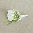 A refined white orchid boutonnière is showcased on a pale, sand-toned woven fabric, its placement slightly diagonal as though it has just been set down on a tailor's table above Highgate High Street. The orchid itself is immaculate, with four or five generous petals shaped like smooth, folded silk, their bright white softened by a whisper of cream at the centre. The throat of the bloom glows with a subtle yellow hue, drawing the eye inward toward the flower's intricate core. Surrounding this central orchid are airy clusters of baby's breath, each stem branching into dozens of minuscule white blossoms, creating a misty, cloud-like collar that introduces texture without overwhelming the design. Slender, flexible strips of fresh green foliage arch out from beneath the orchid, their fine lines adding energy and contrast, reminiscent of grasses brushing along the paths of nearby Hampstead Heath. The stems at the base are neatly pulled together and wrapped in a smooth, shining band of white satin ribbon, tailored into a slim handle that lies flush against the fabric surface. Just above the midpoint of the wrapping, a dainty satin bow is tied, its symmetrical loops adding a final note of ceremony and intention. The linen-style background, with its subtle weave and warm beige colour, offers a quiet, natural canvas that lets the pure whites and greens stand out in calm relief. Soft, diffused lighting falls over the boutonnière, highlighting the satin's gentle sheen and the orchid's velvety texture, creating an atmosphere of poised anticipation, perfectly in tune with a Highgate wedding or civil ceremony where small details carry deep emotion.