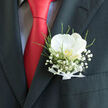 Here, a refined wedding boutonnière is pictured resting in the pocket of a dark, finely cut suit jacket, perfectly complemented by the rich streak of a red tie. The focal point is a single white orchid, its petals smooth as silk and gently cupped, suggesting both delicacy and strength. A soft, creamy glow radiates from the flower, with a whisper of golden yellow at its centre that adds subtle warmth and dimension. Encasing this central bloom is a cloud of baby's breath, the minuscule white flowers clustered so closely that they form an airy, lace-like frame around the orchid. Slim sprigs of greenery and fine tendrils of fern thread through the design, introducing natural texture and hints of fresh green that recall the tranquil foliage of Highgate Wood or the gardens of nearby Waterlow Park. The stems are carefully bound with a narrow white ribbon, wrapped smoothly and secured in a discreet knot, demonstrating a florist's practiced precision and attention to every small detail. Against the matte charcoal of the suit, the flowers seem to glow, their brightness further accentuated by the bold red tie, which provides a lively contrast and suggests a joyful, celebratory mood. This boutonnière feels tailor-made for a groom or honoured guest at a Highgate wedding, perhaps before a ceremony in a local church or a reception in one of the village's characterful venues. The composition conveys a mood of thoughtful elegance and intimate romance, the kind of understated yet meaningful floral touch that Flowers Highgate might prepare for couples who want their day to feel both timeless and deeply personal, rooted in the charm of their local neighbourhood.