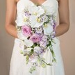 Within a softly blurred, creamy-beige background inspired by the warm tones of Highgate's hillside homes, this richly detailed image frames a bride's mid-torso and elegant strapless dress, which features the delicate texture of lace. Her gently curved arms highlight the radiant cascade of a bridal bouquet, a creation bearing the hallmarks of Highgate's expert florists. At its heart, broad-petalled white phalaenopsis orchids steal the scene, their plush faces glowing with subtle yellow and evoking the serenity of Kestrel Court gardens. Surrounding the orchids, layers of powder-pink garden roses and playful sprigs of lavender and purple-hued lisianthus bring movement and depth, their ruffled petals edged in darker violet-details that speak to the wild, romantic spirit of North London's green spaces. Wispy clusters of white baby's breath drift through the bouquet, while a minimal touch of fresh foliage provides gentle definition. The arrangement tumbles gracefully downwards in a classic cascading silhouette, blending contemporary luxury with timeless tradition. The bouquet's sophisticated harmony and fresh, locally sourced flowers capture the essence of special moments celebrated in Highgate, making this an inspired floral choice for any bride wishing to evoke romance and refined artistry on her wedding day.