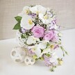 A sumptuous cascading bouquet is artfully arranged against a softly glowing, off-white background, capturing the essence of a bridal design ready to be carried down the aisle of a church or registry office in Highgate. The arrangement forms a refined teardrop shape, with the fullest concentration of blooms near the top, gently tapering into a flowing tail of flowers that drapes downward. Dominating the upper section are several large white orchids, their petals wide and smooth like silk, each blossom featuring a detailed centre touched with warm yellow and gold that brings subtle radiance to the composition. Nestled among these are clusters of roses in tender blush pink and muted lavender, some half-open and others fully unfurled, revealing spirals of velvety petals that give a sense of depth and romantic softness. Between the main flowers, delicate sprays of white baby's breath are woven like fine netting, filling the bouquet with dozens of tiny white dots that add lightness and cohesion. Smaller lilac and white blooms enhance the colour story and soften the transition into the trailing portion of the bouquet. From the lower edge, cascading stems of pale purple blossoms extend gracefully, creating a sense of movement as if the flowers are gently flowing with each step the bride takes. Hints of fresh green foliage appear sparingly, adding definition and a natural, garden-inspired touch without interrupting the pastel scheme. Supporting the bouquet is a white, textured base or decorative stand, while several small, white, cut-out ornamental spheres sit nearby on the pale surface, their ornate patterns echoing the intricacy of lace. The entire composition feels airy, luxurious, and timeless, perfect for wedding photography, ceremony decor, or inspiration for couples planning celebrations in Highgate.