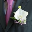 A detailed close-up shows a groom's charcoal suit jacket in soft, even lighting, the camera lingering on a single white orchid boutonniere pinned neatly to the left lapel. The fabric of the suit is smooth and finely woven, almost matte, which makes the satiny, deep magenta tie and light grey patterned shirt beneath it glow subtly by contrast. At the centre of the image, a pristine Phalaenopsis orchid opens its broad, sculptural petals in creamy white, gently cradling a yellow and soft orange throat that catches the light like a tiny sunrise. Around this focal bloom, delicate sprigs of pale lilac flowers and airy white gypsophila cluster together, their tiny blossoms suggesting a miniature wild meadow that has been carefully tamed for formalwear. Deep green leaves peek from behind the petals, adding depth and a natural frame. Just above the main orchid, a single pale green bud stands poised, hinting at new life and the unfolding of the day ahead. The stems are wrapped and finished with a slim lavender satin ribbon tied into an intricate bow, echoing the purple tones of the tie and bringing the whole composition into quiet harmony. The mood is composed and romantic, the kind of understated elegance often chosen for weddings in Highgate churches or intimate ceremonies near Waterlow Park, where a groom might choose this fresh, delicately scented boutonniere to mark a moment of commitment with calm confidence and a touch of modern refinement.