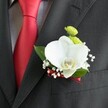 A close-up photograph focuses on a single wedding boutonniere pinned neatly to the lapel of a dark charcoal grey suit jacket, its fine woven fabric softly catching the light. At the centre of the design sits a flawless white Phalaenopsis orchid, its wide, velvety petals unfolding like wings and framing a delicate yellow and white lip that draws the eye. Tucked just above the orchid are two compact, lime-green button chrysanthemum blooms, their rounded forms and many tiny petals adding playful texture and a burst of fresh colour. Around them, wisps of white baby's breath create a cloud of tiny blossoms, giving the boutonniere a light, romantic halo that feels perfect for a Highgate wedding ceremony. A slim deep red satin ribbon wraps the stems at the base, its sheen echoing the rich red satin tie that runs vertically to the left, set against a pale grey and white checked dress shirt. The contrast between the crisp shirt, the vivid tie, and the calm white orchid suggests a groom or groomsman dressed for an elegant celebration at a local Highgate venue, perhaps near Waterlow Park or along the leafy streets by Highgate Village. The lighting is gentle but clear, revealing the smooth orchid petals, the slightly glossy green leaves behind it, and the careful stitching of the suit's lapel and pocket. Every element in the image feels intentional and balanced, from the clean lines of the tailoring to the curved petals of the orchid, capturing the quiet, polished confidence of a thoughtfully chosen floral detail on a special day.