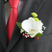 This image showcases a single, thoughtfully composed boutonnière pinned to the lapel of a dark, well-fitted suit jacket, the fabric providing a rich backdrop for the flowers' freshness. At the heart of the arrangement is a white orchid in full bloom, its broad petals unfurling with a smooth, almost sculpted quality. The surface appears velvety yet luminous, catching the ambient light as if in a softly lit Highgate venue. Within the centre of the orchid, a hint of buttery yellow and fine detail marks the throat, giving the bloom a living, breathing presence. Just behind and slightly above, two bright lime-green button chrysanthemums add shape and vibrancy, their rounded heads tightly packed with short petals that create a playful, tactile texture. Light, feathery stems of baby's breath weave around the central flowers, each tiny white blossom spaced along the stem like delicate pearls, softening the outline of the boutonnière and introducing an airy, romantic feel. A few carefully trimmed leaves in a deep green tone cradle the flowers from below, giving structure and contrast. At the base, the stems are neatly wrapped in a narrow, satin red ribbon that coordinates perfectly with the sheen of the red tie visible beneath, binding the look together in a quiet thread of colour. The composition feels intimate and understated, ideal for a groom or groomsman walking through Waterlow Park for portraits or arriving at a ceremony near Highgate Village. The overall mood is sophisticated and serene, a small accent that still speaks clearly of celebration, style, and the craftsmanship of a skilled local florist.