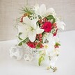 A stunning floral arrangement is presented as a centerpiece, resting on a crisp white surface against a softly textured, warm beige backdrop that could easily belong to a stylish Highgate restaurant or private dining room. The flowers rise from a low, round white container crafted in an open, basket-like weave, its lattice pattern creating an airy, modern base with a handmade feel. Dominating the bouquet are several large white Oriental lilies, their petals spread wide in full bloom, gently curling back to reveal pronounced stamens and soft hints of pollen that lend authenticity and natural charm. Between these impressive lilies, rich red roses introduce deep, velvety tones; some roses are snugly budded, while others are beginning to unfurl into lush, romantic blossoms, bringing a sense of layered depth. Delicate white baby's breath is threaded throughout, its tiny clustered blooms forming a fine, cloud-like mist around the larger flowers. The greens are bright and feather-light, likely fern-like foliage, drawing the eye around the composition and preventing the design from feeling heavy, much like the airy greenery surrounding Highgate's garden squares. Small, glossy clusters of red hypericum berries add playful, festive accents, catching light and offering a subtle seasonal feel. From the right-hand side, a long, arching stem of white phalaenopsis orchids cascades elegantly down, showcasing several open flowers with creamy-white petals and soft yellow-and-pink centers, accompanied by a few unopened buds that promise ongoing beauty. On the left of the arrangement, three white woven spheres lie on the tabletop, echoing the container's texture and rounding out the scene with a minimal, contemporary touch. This harmonious blend of lilies, roses, orchids, baby's breath, and berries creates a luxurious mood, perfect for wedding tables, anniversary celebrations, or intimate dinner parties around Highgate.