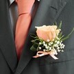 With a quiet air of distinction, this classic buttonhole-made for a Highgate celebration-brings together nature's softness and modern elegance. The image centres on a dark charcoal suit jacket, its fine, almost tactile weave set off by the muted blue checks of the shirt beneath, calling to mind the subtle layering of Highgate's changing seasons. Nestled to the lapel, a single peach rose glows-petals smooth and gently curled, hinting at both romance and fresh beginnings. Frothy clouds of baby's breath wrap delicately around the rose, their white blooms light as mist in the morning air near Highgate Wood. Wisps of green, both slender and broader, sweep behind the arrangement, lending a sense of freshness and lively movement. A neat peach satin ribbon, its gentle sheen reflecting the coral shades of the wearer's tie, finishes the ensemble with polished restraint. Perfect for weddings, proms, or intimate events in the community, this boutonniere is a tribute to artistry and heartfelt local tradition-meant to rest close to the heart and linger in the memory long after the day ends.