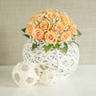 An intimate dome of peach and apricot roses is arranged with the kind of careful restraint you expect from a neighbourhood florist, the blossoms forming a lush, buttery crown. Wisps of white gypsophila drift through the roses like tiny constellations, softening edges and adding a feathery lightness that lifts the whole design. The flowers sit upon a white woven spherical basket, its organic criss-cross of rattan lending a modern, sculptural base that keeps the arrangement visually buoyant. Deep-green foliage frames the lower edge, offering a cool contrast to the warm peach tones and enhancing the bouquet's depth. Warm, directional light-the kind of late-morning glow that filters through the limes along Highgate's roads-gently illuminates each petal, highlighting subtle colour gradations from cream to apricot. You can almost sense the bouquet's scent: a faint, powdery sweetness tempered by fresh green undertones, delicate enough for weddings, anniversaries, or a quiet celebration at home. As someone who arranges flowers for clients across Highgate Village and Hampstead Heath, I choose materials and spacing so the dome reads perfectly from every angle and the gypsophila provides an airy halo rather than clutter. This piece is crafted with longevity and presentation in mind; the woven orb doubles as both vessel and visual punctuation, its shadow play and texture giving the floral mass room to breathe. For customers seeking something serene yet modern, Flowers Highgate often recommends this style for small receptions, mantelpiece displays, or as a bedside bouquet that brings a soft, reassuring presence into the room.