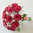 Set against a soft, neutral-toned background, this elegant bridal bouquet radiates classic romance and North London sophistication. The rounded arrangement highlights a wealth of vibrant, velvety red roses, each bloom generous and plush, their rich hues made even more luminous by natural daylight streaming in-a nod to Highgate's leafy lanes and graceful architecture. Delicate clouds of airy white baby's breath are woven throughout the blooms, creating a cloudlike lightness that both softens and elevates the design. Sprigs of dark green leaves edge the bouquet, adding contrast and freshness, while the neatly bundled stems emerge beneath a lavish wrap of wide red satin ribbon stamped with white stripes. The finishing touch-a swirling string of tiny white pearls-lends a sense of refinement, reminiscent of timeless bridal traditions in Highgate's historical venues. The bouquet's polished presentation is matched by the expert local craftsmanship evident in every detail, making it a perfect emblem of love's enduring beauty for same-day delivery across Highgate, Muswell Hill, and surrounding communities.