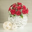 The scene depicts a romantic floral centrepiece styled as if for a small wedding reception in Highgate, where classic red roses meet modern, sculptural design. At the core of the composition is a neat dome of roses in a deep, velvety crimson shade, around a dozen to fifteen blooms gathered close so that their layered petals brush against each other, forming a rich, textured surface. Each rose is fully open yet impeccably shaped, the petals curling inward in tidy spirals that signal freshness and careful handling. Threaded among the roses, clouds of tiny white baby's breath create a gentle contrast, their fine blossoms lighting up the arrangement and evoking the airy quality of tulle or a bridal veil. A few discreet green leaves can be seen at the base, just enough to hint at the natural stems beneath without breaking the disciplined colour story. The bouquet is nestled into a distinctive round vase, bright white and constructed from criss-crossing strands that form an open, woven lattice, giving the impression of a contemporary basket reimagined for a stylish Highgate dining room. This vase stands on a smooth, pale surface, as if on a laid table near Highgate Woods or a townhouse terrace, ready for guests to arrive. To the left of the vase, two decorative spheres made from pale, flat woven strips rest side by side, the larger in front and the smaller just behind, echoing the vase's texture and creating a sense of curated decor. The background is a softly blurred wall in a warm neutral tone, offering subtle texture without distraction. Overall, the image conveys intimacy, elegance, and quiet celebration, perfectly suited to special occasions and romantic dinners throughout Highgate.
