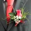 A sharply focused close-up reveals a boutonniere resting on the lapel of a dark charcoal-grey suit jacket, ready for a wedding day in Highgate. At the centre of the design is a single rose bud, coloured in rich fuchsia-pink tones, partially opened so that its velvety petals overlap in a tight, elegant spiral that draws the eye immediately. Clusters of tiny white baby's breath, or gypsophila, surround the rose, creating a delicate halo of blossoms that soften the contrast between flower and fabric. The greenery has been chosen for both texture and shape: smooth, broad leaves with a gentle sheen frame the rose, while finer, feathery sprays add height and a sense of natural, airy movement, like foliage rustling in the breeze near Waterlow Park. At the base of the boutonniere, a bright red satin ribbon is tied into a meticulous bow, its glossy finish beautifully mirroring the bold red silk tie visible beneath the jacket. The tie is sleek and plain, lying flat over a subtly patterned grey dress shirt that adds understated sophistication. The fabric of the suit appears to be a high-quality wool blend, finely woven and soft to the touch, contributing to the overall impression of careful preparation and polished style. Gentle lighting brings out the rich colours and varied textures without harsh shadows, allowing the boutonniere, tie, and lapel to harmonise visually. This image captures a moment of anticipation, the kind of elegant finishing touch a Highgate groom or guest might wear with quiet pride as he heads towards a ceremony or celebration in the heart of the neighbourhood.