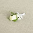 A single ivory rose boutonniere lies diagonally across a softly textured, light beige fabric that looks like natural linen, its fine crosshatch weave catching the gentle light the way table linens might in a Highgate Village restaurant. At the heart of the design is a tightly furled white rosebud, its velvety petals creamy and smooth, with a faint blush of green at the outer edges where the sepals cradle the bloom. Around the rose, clusters of delicate white baby's breath form a misty halo, each tiny flower like a dot of snow, adding airiness and softness to the compact arrangement. Slender, deep green leaves and a few feathery sprigs of foliage extend just beyond the petals, giving a fresh, garden-picked feel that evokes a stroll through nearby Waterlow Park in early spring. The stems are neatly gathered and wrapped in pristine white satin ribbon, the fabric pulled snug and even by a florist's practiced hands, then finished with a small, symmetrical bow positioned just beneath the rose. The satin catches the light with a subtle sheen, echoing the gentle glow on the petal edges. Soft, diffused lighting casts minimal shadows, highlighting the pure white tones and lush greens without distraction, creating a calm, romantic mood ideal for a groom's lapel. The overall impression is of timeless, understated elegance-an intimate piece of wedding floristry that would suit a ceremony in one of Highgate's historic churches or a refined celebration at a local venue, capturing the quiet excitement and promise of the day in a single, carefully crafted floral detail.
