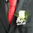 This close-up image reveals a classic ivory rose boutonniere against the dark charcoal of a well-tailored suit, the contrast drawing your attention immediately to the delicate artistry of the flowers. The rose is perfectly formed, its creamy white petals arranged in a natural spiral, neither too tight nor overly open, holding that ideal moment just before full bloom. Around the base, sprays of white baby's breath create a soft halo of tiny blossoms, like a fine mist drifting across the arrangement. Each minuscule flower contributes to a sense of lightness and grace, cushioning the rose with a veil of texture. Slim, glossy-green leaves and slender fern fronds are threaded thoughtfully through the design, adding depth, movement, and a gentle nod to the leafy streets and gardens of Highgate, from Waterlow Park to the edges of Hampstead Heath. The stems are expertly wrapped and finished with a narrow white ribbon bow that rests just above the suit pocket, echoing the rose's purity while adding a subtle, traditional touch. The charcoal suit fabric, crisply cut and smooth, forms a strong frame that allows every floral detail to be clearly seen and appreciated. There is an air of anticipation and quiet pride in the way the boutonniere sits, as if moments before a ceremony at a registry office near Highgate Village or a reception in a nearby townhouse. The overall mood is intimate and refined, perfectly suited to weddings, formal celebrations, or milestone events where understated style and heartfelt sentiment are at the forefront.