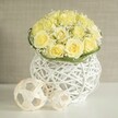 Captured in gentle, diffused light, this floral centerpiece conveys the elegant calm of a Highgate wedding table set for an intimate gathering. At the heart of the composition lies a generous dome of soft yellow roses, the blossoms arranged closely so their petals overlap and intertwine, forming a plush, velvety mound. The colour is a sophisticated creamy yellow, not too bright, lending the design a warm yet tranquil presence. Interspersed among the roses are delicate stems of white baby's breath, their small, cloud-like florets appearing in little clusters that soften the structure and introduce a romantic, airy quality. A slim collar of fresh green foliage wraps neatly around the base of the bouquet, acting as a graceful border that frames the flowers and gently leads the eye toward the vessel. The vase is a rounded white piece created from interwoven strands, almost like a contemporary wicker basket, with open spaces between the lines that let light pass through and create intriguing patterns on the white tabletop below. Resting nearby are two pale woven spheres, their looping, crisscrossing strips forming hollow orbs that mirror the vase's texture and add a sculptural element to the scene. The background is softly blurred in shades of warm beige or stone, evoking the quiet interiors of Highgate homes or a small local venue prepared for a celebration, without any sharp details to distract from the flowers. Overall, the image suggests refined joy and thoughtful craftsmanship, making this arrangement perfect for wedding receptions, anniversary dinners, or special gatherings overlooking the greenery of Highgate Wood or Waterlow Park.