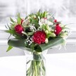 A rich, romantic bouquet stands in a clear cylindrical glass vase, as if freshly set on a sunlit Highgate windowsill. The waterline reveals neatly trimmed green stems, aligned with care to show their natural strength and freshness. Above, a lush crown of velvety deep red roses and a large crimson carnation commands attention, each petal ruffled and softly unfurling, suggesting passion, affection, and timeless love. Around these bold red blooms, creamy white flowers open gently: white alstroemeria with slender petals and delicate speckles, plump white chrysanthemum heads, and several elegant lily buds still tightly closed, promising future blossoms. Fine clusters of white gypsophila-baby's breath-drift between the larger flowers like tiny stars, adding a misty lightness that softens the strong reds. The arrangement is framed by generous, glossy dark green leaves, some broad and tropical, curled into graceful loops around the base to create a structured collar. Eucalyptus-like foliage and pointed green leaves thread through the design, giving layers of texture and shades of green. The bouquet sits on a pristine white surface against a softly blurred, bright background, reminiscent of natural daylight filtering through a Highgate townhouse window. The overall feeling is warm, polished, and welcoming, perfect as a centrepiece for an anniversary dinner, a thoughtful birthday surprise, or a morning delivery to brighten a home office near Waterlow Park. Every element-from the glimmering vase to the interplay of red, white, and green-reflects the attentive craftsmanship of a local florist, presenting a bouquet that radiates quiet celebration and heartfelt sincerity.