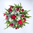 A richly detailed, top-down photograph of a hand-tied bouquet shows a tightly clustered circle of red, white, and green blooms laid out against a pure white background, making every colour and texture stand out. Deep crimson roses sit at the heart of the arrangement, some still in firm bud, others just beginning to open so you can almost feel the velvety spiral of their petals, radiating romance and quiet sophistication. Around them, ruffled fuchsia and ruby-red carnations introduce a playful energy, their densely layered petals catching the light and adding a sense of movement, as if they're gently rustling in a Highgate breeze. Nestled between the richer tones, small white spray chrysanthemums and neat, daisy-like heads create crisp points of brightness, echoing the clean freshness of an early morning walk across Hampstead Heath. Tiny white gypsophila, or baby's breath, is threaded delicately through every pocket of space, forming a fine, misty veil that softens the contrasts and adds a dreamy, cloud-like texture. The flowers are cradled in generous greenery: broad, tapered dark green leaves from lilies or alstroemeria give the bouquet structure, while rounded, silvery eucalyptus leaves weave in and out, cooling the palette with their blue-green sheen and suggesting a soothing, herbal scent. The overall form is full, rounded, and carefully balanced, suggesting a bouquet chosen to make an instant impact when delivered to a townhouse near Highgate Village or a cosy flat off Archway Road. The mood is joyful yet refined, perfectly suited to mark an anniversary, celebrate a birthday, or simply send an affectionate thinking of you that feels both modern and timeless.