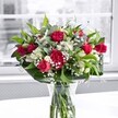 A lush, romantic bouquet of red roses, red carnations, white alstroemeria, and airy baby's breath stands proudly in a clear glass vase on a bright white table, bathed in soft natural light from a nearby window in a Highgate home. The deep crimson roses, some fully open with velvety petals and others still in elegant bud, rise at the heart of the arrangement, giving it a passionate, celebratory feel suited to anniversaries, heartfelt thank-yous, or simply brightening a quiet corner overlooking Waterlow Park. Around them, plump red carnations with finely ruffled petals add volume and a touch of classic charm, their rounded forms creating a rich cushion of colour. Threaded between the reds are crisp white alstroemeria, a mix of tight buds and half-open blooms with trumpet-shaped petals and pale green tips, suggesting beauty that will continue to unfold over the coming days. Sprays of delicate white gypsophila drift through the design like tiny floating stars, softening the contrast between the bolder flowers and catching the faint reflections from the glass vase. The bouquet is framed by layers of glossy deep green foliage and longer, lighter stems that may be eucalyptus, arching gently to give the piece movement and depth. Through the vase, neatly trimmed stems are clearly visible, gathered in fresh, clear water that reinforces the sense of just-arrived freshness you might expect from a local Highgate florist offering same-day delivery. Behind the bouquet, the window view is intentionally blurred and muted in greys, so the reds, whites, and greens appear even more vivid, as if the flowers themselves are the true focus of the room. The overall impression is of a thoughtfully crafted, uplifting arrangement that feels both refined and warmly inviting, perfect for a cosy townhouse near Highgate Village or a modern flat off Archway Road.
