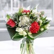 A contemporary glass-vase arrangement is captured in soft natural light, as if placed near a bright window in a Highgate townhouse overlooking Waterlow Park. At the centre, a velvety coral-red rose and a rich crimson carnation provide a dramatic focal point, their layered petals plush and almost touchable. Around them, clusters of delicate white chrysanthemums and tiny daisy-like blooms form a gentle halo, their snowy petals catching every glimmer of light and adding a calm, cloud-like softness. Creamy white lilies, some in tight bud and some just beginning to unfurl, rise gracefully through the bouquet, their long, elegant forms giving height and quiet poise. Broad, glossy emerald-green leaves are carefully folded and wrapped around the base, creating smooth arcs that cradle the flowers and give the whole design a clean, modern silhouette. The clear glass vase reveals a neat grid of stems, held in clear water that sparkles where the daylight touches it, suggesting freshness and careful conditioning in the studio of a local Highgate florist. The overall feeling is one of uplift and gentle celebration, like a calm moment on Highgate High Street after the morning rush. You can almost sense the crisp, leafy fragrance mixed with a light sweetness from the lilies and rose, making this arrangement perfectly suited to marking birthdays, small gatherings, or simply bringing a refined touch of joy to a kitchen table or hallway console in an N6 home.