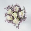 High-angle overhead photograph of a compact hand-tied bouquet where six pristine, creamy white roses sit at the very heart of the design, their petals tightly furled in perfect spirals that look almost porcelain-smooth. Each rose shows a subtle blush of pale green where the sepals meet the stem, adding a fresh, just-cut feeling that any florist in Highgate would recognise from early-morning flower market runs. Around these central blooms, a thick collar of delicate lavender and muted lilac limonium (or statice) creates a soft, feathery ring, the tiny star-shaped flowers forming a cloud-like halo that frames the roses without overwhelming them. The colour contrast is gentle rather than bold: the cool white of the roses glows softly against the powdery purples, suggesting calm, tenderness, and quiet romance. The bouquet is arranged into a neat, rounded dome, as if ready to be carried down the aisle at a small Highgate Village wedding or placed as a graceful gesture of sympathy in a home off Highgate High Street. It rests on a smooth, off-white or very light grey surface-perhaps a clean studio table or a windowsill catching northern light from a Highgate townhouse-so there are no distractions from the flowers themselves. The lighting is even and diffused, with no harsh shadows, allowing every petal edge and every tiny filler bloom to be clearly defined yet softly illuminated. The overall mood is serene and reflective, ideal for marking new beginnings, offering comfort in quieter moments, or adding a refined, understated touch of beauty to an intimate dinner setting near Waterlow Park or on a quiet side street leading up to Highgate Cemetery.