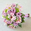 Displayed on a light grey, linen-textured surface, this hand-tied bouquet looks as though it has just been set down between photographs at a Highgate wedding. The soft, neutral background allows the colours to glow gently, especially the luxuriant lavender roses that form the heart of the arrangement. Each rose is in generous bloom, petals layered in soft spirals that evoke scented summer walks along Highgate's tree-lined streets. Between them, pink alstroemeria blooms unfold, their narrow petals touched with blush, cream, and gentle strokes of buttery yellow at the throat, each flower lightly speckled in darker tones for a hint of quiet drama. Bright chartreuse green button chrysanthemums bring an unexpected pop of vibrancy, their compact, round shapes adding both texture and a modern lift to the palette. Threaded through the design are tiny sprays of white baby's breath, like clusters of miniature stars, softening the arrangement and creating a floaty, almost ethereal halo, reminiscent of early morning light over Hampstead Heath just beyond Highgate. At the base, the stems are gathered cleanly and wrapped in a wide strip of pink satin ribbon, its smooth, lustrous surface catching the light in gentle highlights. A single strand of small white pearls winds around the ribbon, adding an elegant bridal accent that feels timeless, like a cherished piece of jewellery. The natural stems extend below, green and woody, giving a freshly cut, garden-inspired character. This bouquet radiates gentle joy and refined beauty, ideal for walking down the aisle in Highgate, gracing a registry table, or marking a significant celebration with soft colour and expert floristry.