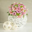 In this serene, close-focus image, a domed pastel bouquet sits gracefully on a pristine white tabletop, bathed in gentle, diffused light reminiscent of a calm afternoon in Highgate Village. The arrangement is lush and rounded, composed primarily of full-bloom roses in delicate lavender and romantic soft pink, their petals layered so densely that they form plush, cloud-like rosettes. Interlaced among them, pale pink alstroemeria lift their graceful, trumpet-shaped flowers, each petal streaked with fine, darker markings that give a sense of depth and movement. To punctuate the softness, bold lime-green button chrysanthemums are tucked between the blooms, their tight, button-like heads bringing a lively, modern contrast and a hint of playful energy. Fine branches of white baby's breath wander through the design, tiny star-like blossoms adding a misty, airy quality that prevents the bouquet from feeling heavy. That lightness is further enhanced by glimpses of dark green foliage at the base, framing the flowers while maintaining the pastel focus. All of this is gathered into a distinctive spherical vase made from white woven wicker or rattan, its open, lattice-like design creating a sculptural cage around the stems and allowing light to filter through. On the tabletop nearby, two smaller decorative spheres crafted from pale, natural woven strips echo the same textural style, adding balance and harmony to the scene. The background is a gently textured neutral wall, likely soft beige or off-white, which recedes quietly so the eye remains on the flowers. The overall feeling is one of refined celebration and contemporary romance, perfectly suited to wedding collections, bridal showers, or elegant home décor for residents across Highgate and the nearby streets leading toward Hampstead Heath.