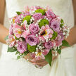 In this image, a hand-tied wedding bouquet has been styled with the quiet sophistication you might expect from a Highgate florist preparing flowers for a ceremony near the village. The centre is dominated by lilac and dusky mauve roses, their petals layered in gentle spirals that open gradually toward the edges, each bloom offering a sense of composure and romance. These cool-toned roses form a soft focal point, evoking calm and tenderness, like the stillness of Highgate Wood just after sunrise. Interlaced among them, delicate gypsophila adds a whisper-light texture, the tiny white blossoms clustering like a fine mist, softening the lines between each flower and giving the bouquet a subtle, airy halo. Bright green button chrysanthemums punctuate the design with flashes of lively chartreuse, their compact, rounded heads and slightly ruffled surfaces providing both contrast and touchable texture. Around this ensemble, pink alstroemeria flow outward in graceful arcs, petals lightly streaked and brushed with warmer hues, adding a sense of motion that gives the bouquet a natural, garden-picked feel while still remaining precise and polished. A base of fresh green foliage, in varied tones of emerald and leaf green, frames the composition and supports the flowers, ensuring the overall shape remains balanced and comfortable in the hand. Soft, diffused light illuminates the arrangement, catching the velvety rose petals and the bright centres of the chrysanthemums, creating gentle highlights and shadows that suggest depth and volume. It feels like a bouquet ready to leave a Highgate studio for same-day delivery to a ceremony in a local church or a reception overlooking the city, created with careful attention to how each bloom will look and feel throughout a long, joyful day.