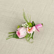 Resting on a softly patterned cloth, this pink rose boutonniere feels like a small work of art thoughtfully crafted for a special day in Highgate. The central rose is a cool blush shade, its petals unfolding in gentle layers that transition from a richer pink in the heart of the bloom to softer, lighter tones at the edges, where they catch the ambient light. The bloom looks freshly cut, with a subtle, velvety sheen that suggests it has just been delivered from a trusted local florist. Closely arranged around the rose are pastel alstroemeria florets, their slender petals gently arched and decorated with fine natural streaks, each blossom holding a touch of yellow at the centre that lends a quiet glow, much like the soft light that filters through the trees in Highgate Wood. White gypsophila clusters float between and around these blooms, adding a lace-like softness and a sense of delicacy. Sprigs of greenery thread through the design, some narrow and fluid, others slightly fuller, giving structure while maintaining an effortlessly natural look. The stems are gathered at the base and wrapped in a narrow pink ribbon, smooth and slightly glossy, keeping everything neat and comfortable to pin against a lapel. The fabric underneath, likely in a neutral tone, provides gentle contrast, highlighting each element's colour and texture. Imagining touching it, one might sense the mix of textures: the rose's velvet, the alstroemeria's silkiness, the gypsophila's lightness, and the crisp foliage. Suitable for a wedding near Highgate Village, a ceremony overlooking the city, or a milestone celebration, this boutonniere speaks of thoughtfulness, romance, and the subtle elegance that suits the character of the area.