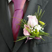 A single wedding boutonnière is pictured in crisp focus, an elegant composition that would feel perfectly at home on a groom's lapel at a ceremony overlooking Highgate Woods. At its heart is a gently cupped pink rose, the shade soft and romantic, with each petal curving inward to create a smooth, spiralling centre. The rose is framed by cool green foliage, including slender, pine-like sprigs and a neatly folded leaf that shapes the outline with the same precision as a well-cut suit. Tiny white gypsophila flowers weave through the greenery, their clustered blooms forming a fine, frothy texture that brings sweetness and a sense of airiness to the piece. Tucked in beside the rose, a crisp green button chrysanthemum adds a vibrant flash of lime, its round, structured form adding a modern, almost graphic note to the arrangement. The stems are bound tidily at the base, suggesting secure fastening to a jacket or tuxedo, the flowers angled so they sit proudly without appearing stiff. Against a dark lapel, the colours come alive: the blush of the rose, the clear white of the gypsophila, the fresh green of the chrysanthemum and foliage. The boutonnière feels both effortless and intentional, like a quiet signature of style chosen with care. One can imagine its subtle fragrance mingling with the air at a Highgate wedding, a prom at a local school, or an intimate celebration along the slopes of Highgate Hill - a small but meaningful floral detail, crafted by a local florist with a practiced eye for balance, proportion, and romance.