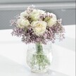 A softly lit close-up shows an elegant bouquet of creamy white roses cradled in a cloud of lavender-purple statice, arranged in a clear crackle-glass vase on a bright white surface. Each rose is captured at that perfect moment of opening, its velvety petals unfurling in gentle spirals, the centres touched with the faintest hint of pale butter-yellow that suggests absolute freshness. The roses sit slightly above the surrounding flowers, like small moons rising from a mist, while the statice forms a delicate halo around them, thousands of tiny papery blooms clustering on slender stems. Their muted lilac tones bring a cool, calming contrast to the warm cream of the roses, and together they create a composition that feels both refined and quietly romantic. The vase itself has an icy, crackled texture that fractures the daylight into soft glimmers along its surface; through the clear water you can see the green stems, neatly trimmed and fanning out to give the bouquet a balanced, rounded shape. The white tabletop beneath reflects some of the light, casting faint, feathered shadows that enhance the sense of airiness and space, while the background falls away into a blurred wash of pale grey. There are no distractions, only the serene beauty of the flowers themselves, as if they have been placed on a windowsill in a Highgate townhouse overlooking Waterlow Park. The mood is tranquil and contemplative, making this arrangement ideal for a gentle birthday surprise, a quiet get-well wish, or simply to bring a moment of stillness and grace into a calm corner of a home or studio near Highgate Village.