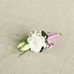 On a neutral, textured fabric that resembles light linen, an exquisite boutonniere lies ready for a celebration, as if placed on a windowsill overlooking Highgate's treetops. At its heart is a fully open lisianthus bloom, creamy white and abundantly ruffled, with petals that fold and ripple like fine, lightweight fabric. The central petals gather in soft layers, creating a rounded, almost cloud-like form that exudes calm and sophistication. Nestled alongside this main flower are two graceful lisianthus buds, their elongated shapes still partially closed, tipped with gentle green tones that speak of new beginnings and unfolding stories. Draped between bloom and buds are airy stems of baby's breath, each one dotted with a constellation of tiny, white blossoms. These add a lace-like veil across the arrangement, enhancing its romantic, almost poetic character, reminiscent of the soft ambience around Pond Square on a quiet afternoon. Slender trails of green foliage extend around the flowers, adding light movement and the freshness of a garden-cut posy. The stems are gathered and carefully wrapped with a bright pink satin ribbon, its smooth, glossy texture catching the light in subtle highlights. The ribbon is finished with a small, precise bow, loops and ends aligned, bringing a playful yet elegant accent of colour that contrasts beautifully with the creamy whites and tranquil greens. This boutonniere is perfectly suited for a groom or guest at a Highgate wedding, a civil ceremony near Waterlow Park, or a special event along Highgate Hill, embodying gentle romance, careful craftsmanship, and the refined charm of a local florist's work.