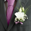 This groom's boutonnière, perfectly positioned on a deep charcoal lapel, combines classic floral elegance with a quiet, contemporary sensibility that suits a Highgate wedding beautifully. The focal point is a single white lisianthus bloom, open and serene, its petal edges lightly frilled and layered so that they seem to fold and curl like soft fabric. In the gentle light, the petals pick up delicate shadows, giving the flower a sculpted, almost porcelain quality. Behind the bloom, two lisianthus buds stretch gently upwards, slender and smooth, suggesting both growth and the couple's shared future. Around them, fine, branching stems of baby's breath create a soft halo, each tiny white flower a pinprick of brightness that calls to mind a veil of mist over Highgate's leafy streets. Slim, fresh green foliage threads through this airy cloud, some pieces arching gracefully, others standing upright, adding an easy, natural rhythm to the composition. At the base, a single deep green leaf is looped and neatly tucked, offering a polished, glossy frame that contrasts with the delicate textures above. The stems are wrapped in a muted mauve ribbon, its satin surface catching light with a subtle sheen. The ribbon is tied into a tidy bow that rests just below the bloom, its gentle lavender tone harmonising with a silk tie or pocket square without ever overpowering the white of the lisianthus. As the groom moves-perhaps stepping out of a car on Highgate High Street or walking into a ceremony near Parliament Hill-the boutonnière appears to shift softly, the petals and baby's breath trembling slightly and releasing the faint suggestion of a clean, floral fragrance. The overall feeling is one of composed joy and thoughtful craftsmanship, a small floral accent that holds deep emotional resonance.
