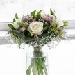 A refined white and green bouquet stands in a simple cylindrical glass vase, placed before a softly glowing white backdrop that suggests a bright Highgate morning filtering through a window. At the heart of the arrangement are several pristine white roses, fully opened yet still velvety and firm, their petals curling outwards in smooth, satiny layers that shade gently from creamy white at the edges to a faint ivory at the centers. Around these focal blooms, a collar of rich emerald foliage fans out, with some broader leaves carefully folded to cradle the roses and give the design a composed, architectural feel. Fine sprigs of white gypsophila, or baby's breath, are threaded delicately between the larger flowers, their tiny cloud-like blossoms catching the light and creating a lacey halo around the central blooms. Slim stems of alstroemeria, some still in bud and others just starting to open, introduce hints of soft mauve and blush-pink, their subtle streaked petals adding depth and a whisper of colour without disturbing the bouquet's overall serenity. Through the clear glass vase, the green stems are clearly visible, neatly aligned and gently bound with a narrow ribbon above the waterline, the fresh water sparkling in the natural light. The whole composition feels calm and balanced, with a rounded, compact silhouette that would be perfectly at home in a quiet Highgate townhouse or by a window overlooking Waterlow Park. It conveys purity, tenderness, and thoughtful elegance, as if prepared by a local florist for a gentle condolence, an intimate wedding, or a quiet moment of reflection, where soft fragrance and understated beauty bring comfort and peace.