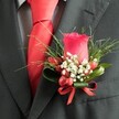 A detailed, high-angle close-up shows a classic wedding boutonnière pinned neatly to the left lapel of a dark charcoal suit jacket, the kind you might see on a groom arriving at a ceremony in Highgate Village. At its heart is a single, perfectly formed deep red rosebud, its velvety petals tightly layered and standing upright with quiet confidence. Around the base of the rose, tiny white blossoms, much like airy clouds of baby's breath, soften the bold colour and add a gentle halo of lightness. Nestled among them are several small, glossy red berries, rounded and plump, echoing the rose's rich crimson tone and giving the arrangement a subtle sense of texture and depth. Lush greenery supports the flowers: fine, grass-like fronds fan out in delicate lines, while broader, smoother dark green leaves hold the composition together like a protective cradle. A narrow red satin ribbon is wrapped and tied into a precise little bow at the stem, its sheen catching the soft indoor light and tying the red elements into a harmonious whole. Beneath the lapel, a bright red silk or satin tie is knotted perfectly, its surface smooth and reflective, reinforcing the rich colour story of the buttonhole. A pale, subtly patterned dress shirt in light grey or white peeks through, perhaps a fine check or houndstooth weave, adding an extra layer of refined texture. The overall mood is one of considered elegance and romantic formality, the sort of carefully coordinated finishing touch a local Highgate florist would prepare for a wedding, prom, or black-tie celebration near Waterlow Park or along Swain's Lane, where every small detail matters.