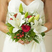 In this image, a luxurious hand-tied bridal bouquet is nestled close to a lace wedding dress, the intricate floral design mirroring the delicate detailing of the fabric. At its heart sits a lush red rose, fully open, each velvety petal curling back gracefully to form a rich, romantic centrepiece. Encircling the rose, generous white lilies stretch their petals outward in smooth, star-like forms, their surfaces catching the light in subtle highlights that make them appear almost luminous. Some lilies are fully open, while others remain as elegant buds, introducing a sense of unfolding time and anticipation. Gentle sprigs of gypsophila drift between the larger blooms like tiny white stars, softening the transitions and adding a misty, romantic feel. Glossy red berries punctuate the bouquet with small bursts of cheerful colour, balanced thoughtfully with rounded, mossy green dianthus heads that bring a fresh, textural contrast and a hint of woodland undergrowth, as though inspired by the greenery around Waterlow Park. Slender, arching grasses and layered foliage form an airy frame around the flowers, lending the bouquet a slightly wild yet carefully controlled silhouette, the kind of aesthetic prized by brides in Highgate who love a natural, garden-inspired look. The stems are neatly gathered, hand-tied to sit perfectly in the bride's hands as she walks through the quiet streets towards her ceremony. The colour palette of creamy white, deep red, and vibrant green is both classic and striking, giving the bouquet a sense of timeless elegance. One can almost picture this arrangement being carried into a historic Highgate church, the soft fragrance of lilies mingling with the emotions of the day-joy, devotion, and the delicate thrill of saying yes to a shared future.