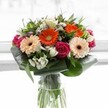Bursting with vibrant colour and structured grace, this bouquet perfectly captures the unique character of Highgate's renowned florists. At the heart, four gerberas-two in exuberant orange and two in understated peach-provide a glowing foundation, their soft petals arranged around dark, expressive discs. Tucked alongside, tightly closed magenta roses offer a romantic note, their velvety texture and deep hue intensifying the sense of occasion. Between them, clusters of snowy alstroemeria and petite white spray roses peek out, their delicate, star-like blooms creating a feeling of abundance and jubilation. Splashes of blush pink and hints of ivory whisper through the greens, inviting feelings of hope and renewal. Broad, glossy leaves wrap the lower bouquet in a sturdy embrace, while finer foliage slips between every stem, giving life and volume to the whole arrangement. All of this is elegantly presented in a tall glass vase, the natural drama of green stems visible through crystal clarity-a sight at home above Highgate Village's cobbles or on a table near Waterlow Park. Drenched in soft natural light, every detail is showcased with pride and expertise, making this bouquet an inspired choice for celebrating milestones, expressing appreciation, or simply sharing a touch of spring with someone cherished in the Highgate community.