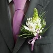 A close-up, high-angle photograph captures the upper torso of a man dressed in refined formalwear, standing in soft natural light that could easily be streaming through a townhouse window off Highgate High Street. He wears a charcoal grey suit jacket with a sharply tailored lapel and clean shoulder line, the dark wool fabric smooth and slightly matte against the lens. Beneath the jacket, a pale dress shirt with a subtle grid pattern peeks through, its cool tones echoed by a perfectly knotted lavender tie that runs in a straight, composed line down his chest. Pinned carefully to the left lapel is a meticulously crafted wedding buttonhole, the clear focal point of the image. At its centre, a tall, pale green spire of unopened buds rises like a tiny spring shoot, surrounded by crisp white Alstroemeria blooms, each petal painted with delicate burgundy streaks and tiny golden stamens. Wisps of white gypsophila cloud around the lilies, lending a soft, almost misty texture. Slender blades of grass and deeper green foliage curve gracefully, framing the flowers and bringing a sense of natural movement that feels at home in nearby Waterlow Park. At the base, a satin lilac ribbon is tied into a neat bow, perfectly matching the tie and drawing the eye to the craftsmanship. The overall mood is elegant, composed, and quietly joyful, the kind of boutonniere a Highgate groom might choose for a wedding in a local church or a stylish celebration nearby, its gentle fragrance and fresh petals marking a moment of loving anticipation.