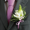 This image shows a finely dressed person's chest and shoulders, framed tightly so that every detail of their formal wear and boutonniere becomes clear, almost tangible. The suit jacket is a deep charcoal grey with a clean, pressed lapel, giving a contemporary yet classic feel, like something you might see at a smart Highgate wedding reception tucked away on a tree-lined side street. Beneath the jacket, a white dress shirt patterned with very small dark checks peeks through, adding a hint of texture without distracting from the main composition. A solid lavender tie falls in a smooth line down the centre, its satin finish catching the light just enough to look refined rather than showy. Pinned to the left lapel is a carefully designed boutonniere crafted from white Peruvian lilies, their petals creamy and soft with fine reddish-brown streaks and golden throats, all arranged to face outward in a gentle arc. Rising from the middle of the flowers, a tall, pale green, conical bud lends a fresh and slightly architectural element, while clusters of airy baby's breath dance around it like a light mist. The arrangement is framed by layered greenery: feathery fronds, narrow spiky blades, and broader, glossy leaves, each one placed so the composition looks both natural and intentional. Around the stems sits a lilac ribbon tied in a small, elegant bow, perfectly matching the tie and bringing a sense of unity to the look. The overall impression is one of cool freshness and quiet anticipation, ideal for a milestone moment, from a prom photo on Highgate Hill to a civil ceremony just a short stroll from the Highgate Underground station.