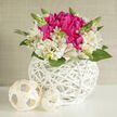 On a smooth, pale surface, a rounded wedding arrangement glows softly, the kind of bouquet that might grace a sunlit table in a Highgate café or a boutique ceremony venue near Highgate Wood. At the centre of the design, vivid fuchsia flowers bloom in lush, ruffled clusters, their petals overlapping generously to form a plush, almost cushion-like core. The colour is bold yet elegant, hinting at passion and joy without overwhelming the senses. Surrounding this vibrant centre is a halo of creamy white alstroemeria, each blossom with slender petals that extend outward like small wings. Fine magenta streaks run along some of the petals, creating a delicate conversation between the soft outer ring and the brighter centre. The bouquet emerges from a perfectly round white wicker basket whose intertwined bands form a light, airy lattice. Every curve of the weave is visible, giving a hand-crafted feel that echoes the artisan shops sprinkled around Highgate Village. Two cream-coloured woven spheres are placed nearby, echoing the texture of the basket and framing the arrangement with quiet balance. Bright green leaves, stems, and a few upright buds peek through the flowers, adding movement and a gentle sense of growth, as if the bouquet has just been gathered from a carefully tended garden. The overall lighting is calm and even, highlighting the smooth petals and the subtle shadows cast by the wicker. This design captures the mood of a refined celebration: perfect as a wedding table centrepiece, a romantic anniversary surprise, or an elegant gift for a Highgate home, where understated luxury and natural beauty are cherished in equal measure.
