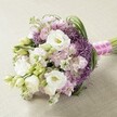 On a pale, textured cloth reminiscent of Highgate's signature Georgian interiors, a lavish bouquet radiates sophistication and a touch of wild English romance. Central to the design are clusters of lush, full-petalled white Lisianthus (Eustoma), their layered ruffles catching the daylight and recalling the refined elegance found in gardens trailing Muswell Hill. Complementing these are blooms in shades of lavender and pastel pink, such as graceful Snapdragons and ruffled Stock, interspersed for gentle contrast. Deep violet Allium spheres punctuate the arrangement with bursts of dynamic, starburst energy, their spiky forms creating lively movement that echoes Hampstead Heath's untamed floral meadows. Slender strands of bright green foliage and unopened buds wind through the rounded, slightly elongated bouquet, adding freshness and a touch of the outdoors. Stems are elegantly bundled with a pastel pink ribbon, striped subtly in white and finished with delicate white pearls-a nod to graceful Highgate wedding traditions. With soft, even light illuminating the intricate interplay of colour and texture, this bouquet radiates joy and serene celebration, making it perfect as a bridal bouquet, a sophisticated centrepiece, or an emblem of Highgate's enduring commitment to classic floral art.