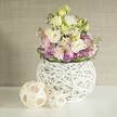 A scene of sophisticated calm, this Highgate-inspired still life features a lush, handcrafted bouquet that radiates understated grandeur. Central to the composition, creamy white and pale yellow lisianthus-each petal finely ruffled-are paired with vertical pink stock blossoms to create a multidimensional sense of height and grace. At the base, a dense scattering of rich purple filler flowers, likely allium or aster, brings depth and shadow, while sprigs of fresh green foliage gently cradle the design. The arrangement rests in a spherical, open-work white vase, its modern texture suggesting a weave of painted rattan reminiscent of the area's love for artisanal, individual style. Alongside, two decorative rattan spheres echo the craftsmanship of the vase, their airy patterns catching the soft natural light that pervades the room. The background-a muted beige wall with subtle, fabric-like texture-completes the tableau, offering a serene, neutral stage for the bouquet's colours to shine. The uplifting ambiance calls to mind tranquil mornings in Highgate Village or an elegant afternoon gathering at Waterlow Park. This arrangement, suitable for wedding receptions or home entertaining across Highgate, conveys a message of renewal, quiet joy, and the season's fresh promises, shaped by the knowledgeable hands of a true local florist.
