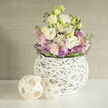 Soft spring light seems to gather around this serene bouquet, a rounded and generous composition that reads both contemporary and comforting. Creamy white and pale buttery yellow Lisianthus (Eustoma) form the principal blooms, their many-layered petals unfolding like silk against the blush clusters of pink Stock; small, spiky purple Allium blossoms punctuate the arrangement with playful geometry while tender green foliage washes through and lifts the palette. The bouquet is tucked into a distinctive spherical white vase made from a web of slim white branches - a sculptural lattice that lets you glimpse stems and water, marrying artisanal craft with botanical clarity. Two natural-tone woven orbs rest to the left on the same smooth white shelf, their rattan-like weave echoing the vase and providing a soft counterpoint of texture. The background is a soft, neutral greige with subtle texture-linen or grasscloth-blurred to keep attention on the flowers. Imagine this piece arriving in a Highgate parlour or brightening a kitchen table near Archway Road; it reads as a thoughtful gift or a calm centrepiece for a bedside or small gathering. The scent would be discreet-light musk with a green, floral sweetness-and the overall mood is one of refined ease. As a local florist familiar with the neighbourhood markets around Highgate, I choose these pastel tones and tactile materials to convey warmth, restraint, and the quiet joy of fresh blooms in an urban home.