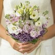 There is a gentle poetry within this Highgate bridal bouquet, each element chosen to evoke the tranquil elegance of its surroundings. Ruffled white lisianthus unfold in creamy layers, their centers touched softly by a dash of green, while haloed among them are clusters of pastel stock in pink and pale lavender, scenting the air much like spring breezes through Highgate Wood. At the bouquet's base, a crown of spiky, star-shaped purple allium offers bold contrast and modern structure, a nod to the area's beloved floral borders. Slender loops of glossy bear grass wrap the midsection, sculpting the composition and lending a flourish of movement and artistry. The bridal hands below are manicured in understated blush, cradling the bouquet close, set against a blurred glimpse of flowing ivory-suggestive of joyful anticipation on Southwood Lane. This arrangement, made with a confident eye and loving touch, radiates hope, romance, and the freshness of local blooms at their peak-a perfect reflection of a Highgate celebration, ready to mark a wedding day or special event with unforgettable style and fragrance.