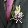 A close-up wedding portrait focuses on the upper body of a man in a sharply tailored charcoal suit, his lapel framed in soft, natural daylight, somewhere that feels like a smart Highgate townhouse or a reception near Waterlow Park. The suit fabric has a fine, almost brushed texture, its dark tone setting off the crisp, pale dress shirt beneath, where a subtle grid pattern quietly catches the light. At the centre of the image, a bold magenta silk tie glows with a gentle sheen, the knot perfectly shaped and resting against the shirt with an easy confidence. Pinned carefully to the left lapel is a delicate, handcrafted buttonhole: slender, closed buds of creamy white flowers rise at the top, while beneath them sit ruffled petals in pastel pink and soft lavender, reminiscent of sweet peas or scented stock gathered from a North London flower market. Tiny clouds of white gypsophila are threaded through the arrangement, giving it a light, airy halo, while a few deeper purple florets draw the eye inward and add quiet depth. Polished dark green leaves support the blooms, their broad, glossy surfaces contrasting with finer, feathery fronds of bright foliage that arc softly outwards, like the edge of a hedgerow in nearby Highgate Woods. At the base, a neat bow of lavender satin ribbon cinches the stems with a satiny glint that echoes the floral tones and complements the tie without competing. The overall impression is one of modern elegance and understated romance: a groom or cherished guest ready for a ceremony in Highgate, his buttonhole a small yet meaningful flourish that speaks of fresh flowers, careful craftsmanship, and the excitement of a day filled with celebration.