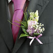 This image shows a carefully crafted boutonnière resting against the lapel of a dark, tailored suit, its soft hues and intricate textures forming a delicate point of focus. At the heart of the design sit pale lavender blossoms, their petals slightly ruffled and velvety, layered together like folded silk. Around them, small white filler flowers-dainty gypsophila and similar frothy blooms-spill out in tiny clusters, creating a halo of lightness that contrasts gently with the depth of the suit fabric. Glossy green leaves curve protectively behind the flowers, their surfaces smooth and reflective, as if they had just been misted with water in a Highgate flower studio before the event. A slender, translucent ribbon in a gentle lavender shade is wrapped around the stems, tied neatly and pinned close so that no loose ends disturb the clean lines of the lapel. The tie beneath, in a vivid magenta tone, adds a punch of colour that echoes through the pastel flowers and makes the lilacs appear even softer by comparison. The composition feels intimate and intentional, like a small promise pinned close to the heart-ideal for a wedding ceremony taking place in one of Highgate's historic churches or a civil celebration overlooking the greenery of Parliament Hill. Every element speaks of care: stems cut to the right length, foliage trimmed to keep the shape light, and blooms chosen at just the right stage of opening. It is a subtle emblem of romance and elegance, an accent designed to complement rather than overpower, quietly enhancing the wearer's presence on a meaningful day.