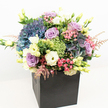 Luxurious and visually captivating, this bouquet bursts with colour and life, reflecting the unique character of Highgate's tree-lined avenues. Expertly arranged in a modern matte black square container, the composition is dense and rounded, structured to showcase each bloom's radiance from every angle. Prominent hydrangeas in multi-toned indigo, blue, and periwinkle anchor the piece, their generous clusters suggesting the lushness of Highgate's gardens. Interlaced within are soft lavender and dusky mauve roses, their petals gradually unfolding, set alongside trumpet-shaped lisianthus in creamy white and gentle yellow, which lend a touch of classic luxury. Vibrant, lime-green 'Green Trick' dianthus and delicate sprays of pale pink astilbe provide playful texture and contrasting pops of colour, their forms reminiscent of the wild blooms trailing along Highgate Wood footpaths. Shiny and matte green leaves, along with emerging buds, weave through the bouquet, enhancing its full, garden-inspired presence. The black container acts as a striking foil to the vivid florals, echoing the area's blend of tradition and modernity. Bathed in bright, even light, the palette of purples, blues, pinks, creams, and lively greens feels celebratory and uplifting-befitting a special Highgate celebration, heartfelt thank you, or tribute. Perfect as a dramatic centrepiece or a luxury delivery to Highgate, this arrangement transforms local flowers into art for the home or workplace.