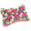 Viewed from a gentle, slightly elevated oblique angle against a clean white backdrop, this pillow-shaped floral tribute reads like a soft, tactile landscape of colour and texture. Densely packed across the rectangular 'cushion' are velvety hot pink roses, some petals still curled into tight buds while others open into plush rosettes, their scent rich and reassuring. Scattered between them are pale blush-pink carnations whose ruffled edges add a delicate, feathery rhythm; small sprays of tiny pink filler blooms provide a whisperlike playback of detail. Bright lemon-yellow ranunculus punctuate the surface with sunny, cup-shaped flashes, while deep purple lisianthus lends cool depth and a graceful, bell-shaped contrast. Architecturally sharp and silver-blue in tone, eryngium thistles interrupt the softness with a striking spiky note, like memories that stand out in relief. A generous layer of rounded, silvery-green eucalyptus and mixed deep-green foliage forms the visual underlay, their matte leaves catching soft studio light and framing the palette. The cushion's perimeter is defined by a ruffled, pleated red satin ribbon - a rich, deep-red border that nests and elevates the floral tapestry and keeps the shape elegant and resolved. The overall mood is one of comforting celebration rather than sombre austerity: lively hues of fuchsia, violet, lemon and blush combine to honour a life with warmth and affection. Handcrafted with careful symmetry and layered textures, this memorial pillow would bring a gentle, fragrant presence to a Highgate chapel, a graveside at the local cemetery, or a quiet family home, offering consolation and a sense of tender remembrance.